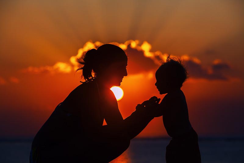 Silhouette of happy beautiful mother smiling to joyful baby boy on sea beach on background of sunset sky with clouds. Motherhood family lifestyle and summer vacation with child on tropical resort