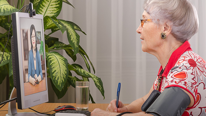 elderly woman is measuring blood pressure by herself during virtual doctor visit. Glasses senior woman sitting opposite monitor. On the screen telehealth doc is consulting her. Side shot