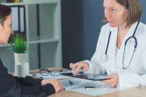 female doctor consulting with a patient at a desk in an office with a serious expression holding a tablet