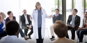 a female doctor addressing other healthcare professionals sitting around her in a circle