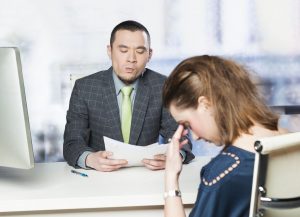 an interviewer looking skeptically at a resume and an embarrassed woman sitting on the other side of the table