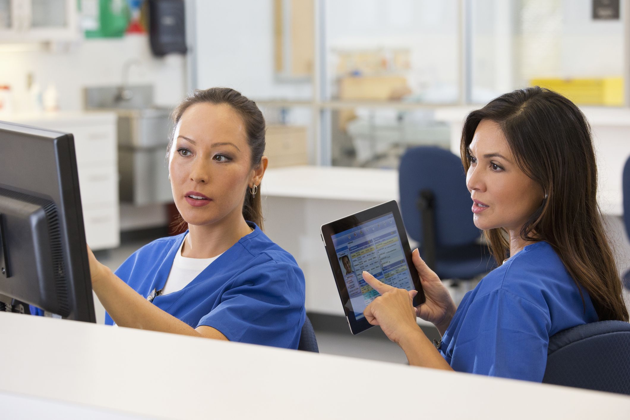 two female healthcare professionals at a nurse's station looking at a computer one is holding a tablet