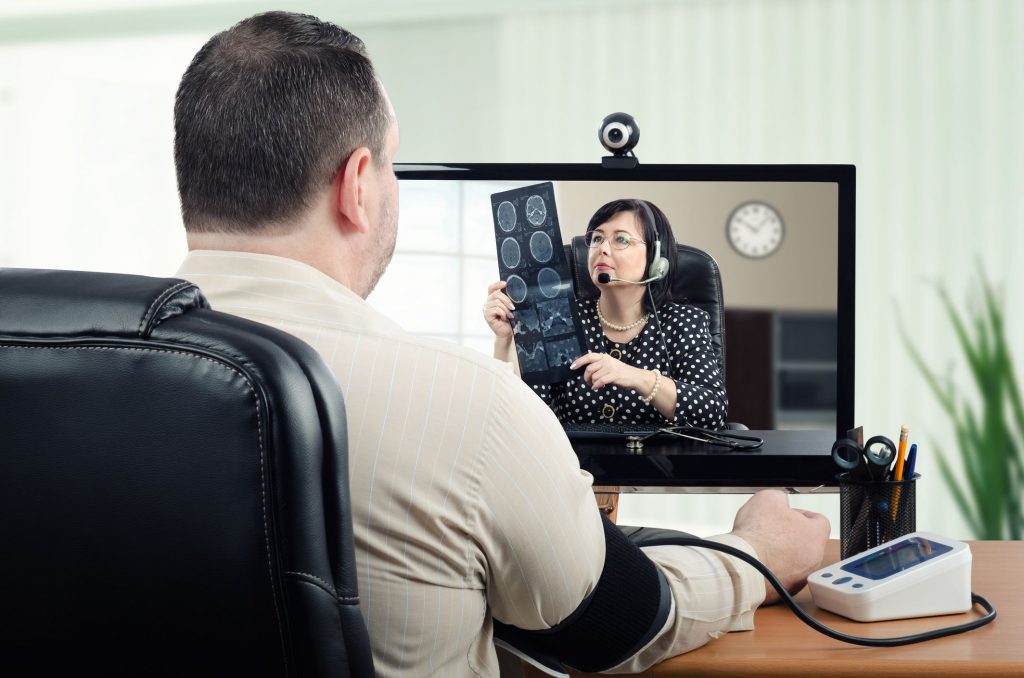 a doctor looking at cat scans on a computer screen having a telehealth appointment with a man facing the screen taking his blood pressure