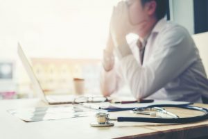 stethoscope on a table in the foreground with a male doctor out of focus at a computer looking anxious