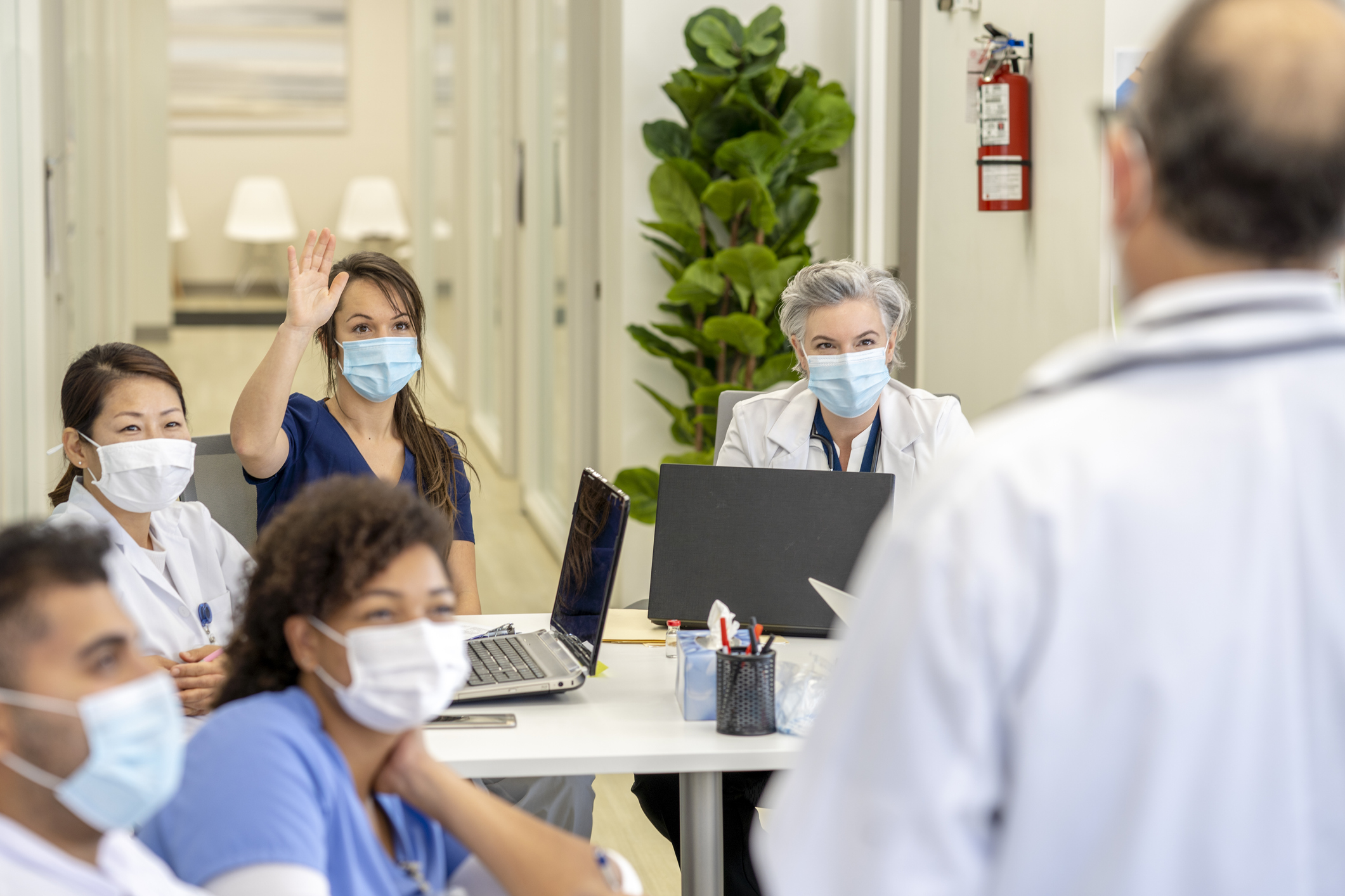 Healthcare professionals meeting together, with a speaker at the front of the room, his back to the camera. Everyone is wearing a face mask to prevent the spread of germs.