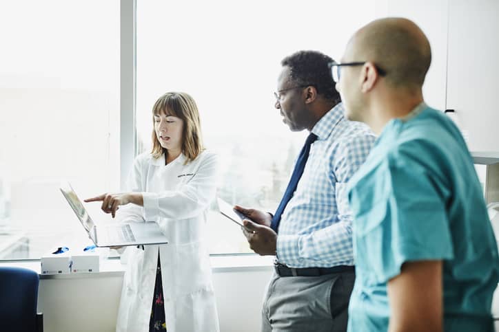 health information manager in lab coat shows health information on laptop computer to two male colleagues