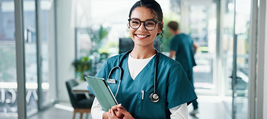 Smiling healthcare professional wearing teal scrubs and a stethoscope, holding a clipboard in a bright medical facility hallway.