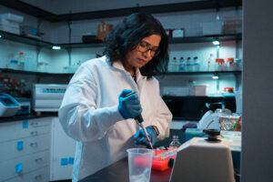 Scientist wearing a white lab coat and blue gloves uses a pipette while working with samples in a laboratory.