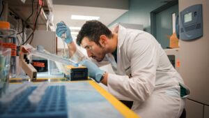 A scientist in a white lab coat and blue gloves examines a laboratory sample using a pipette at a workbench.